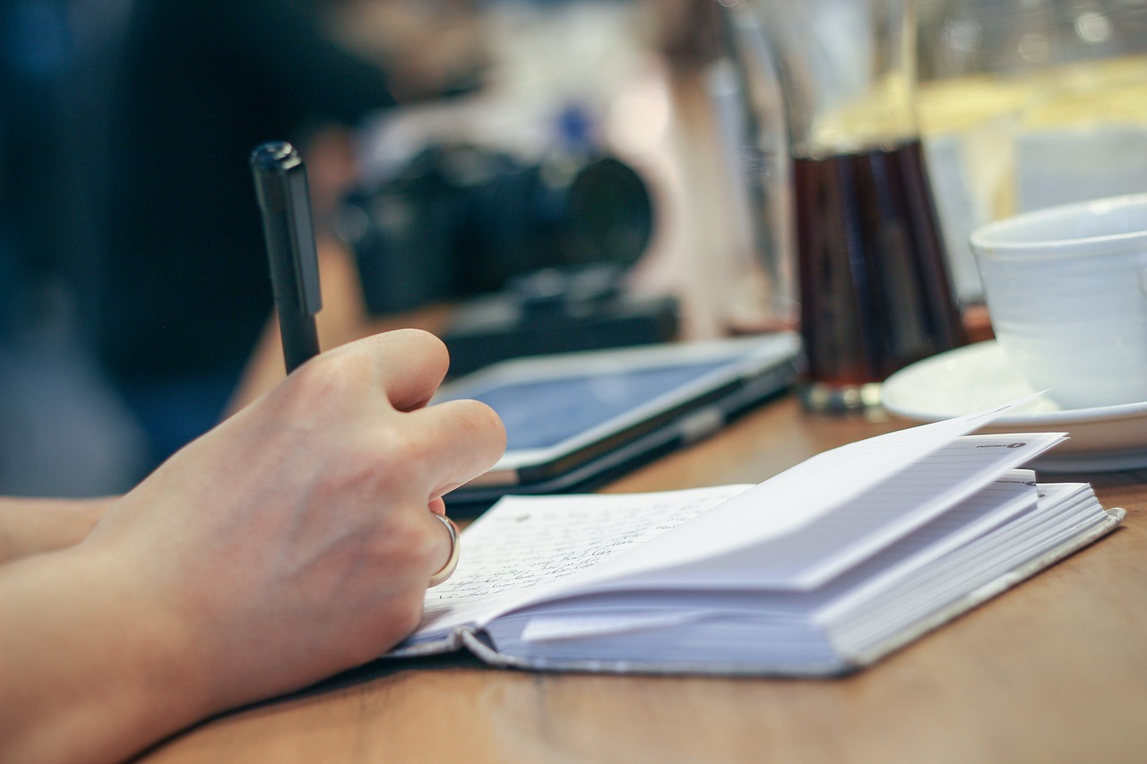 Hand writing translation notes in a notebook at a desk, pen moving across open pages